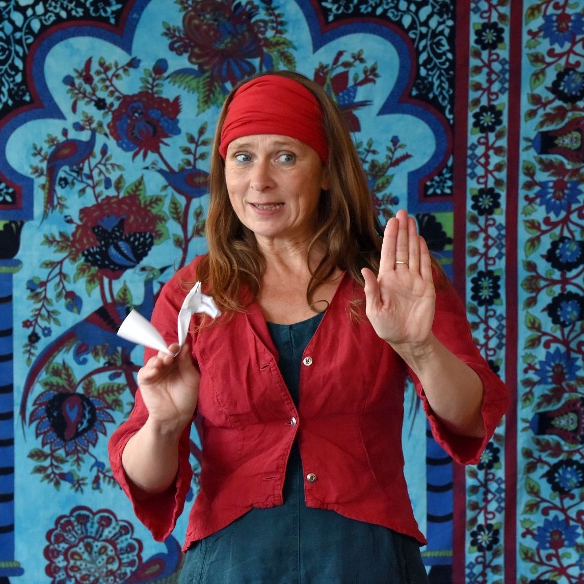 Eine Frau mit einer roten Jacke und einem roten Stirnband steht vor einem bunten Wandteppich