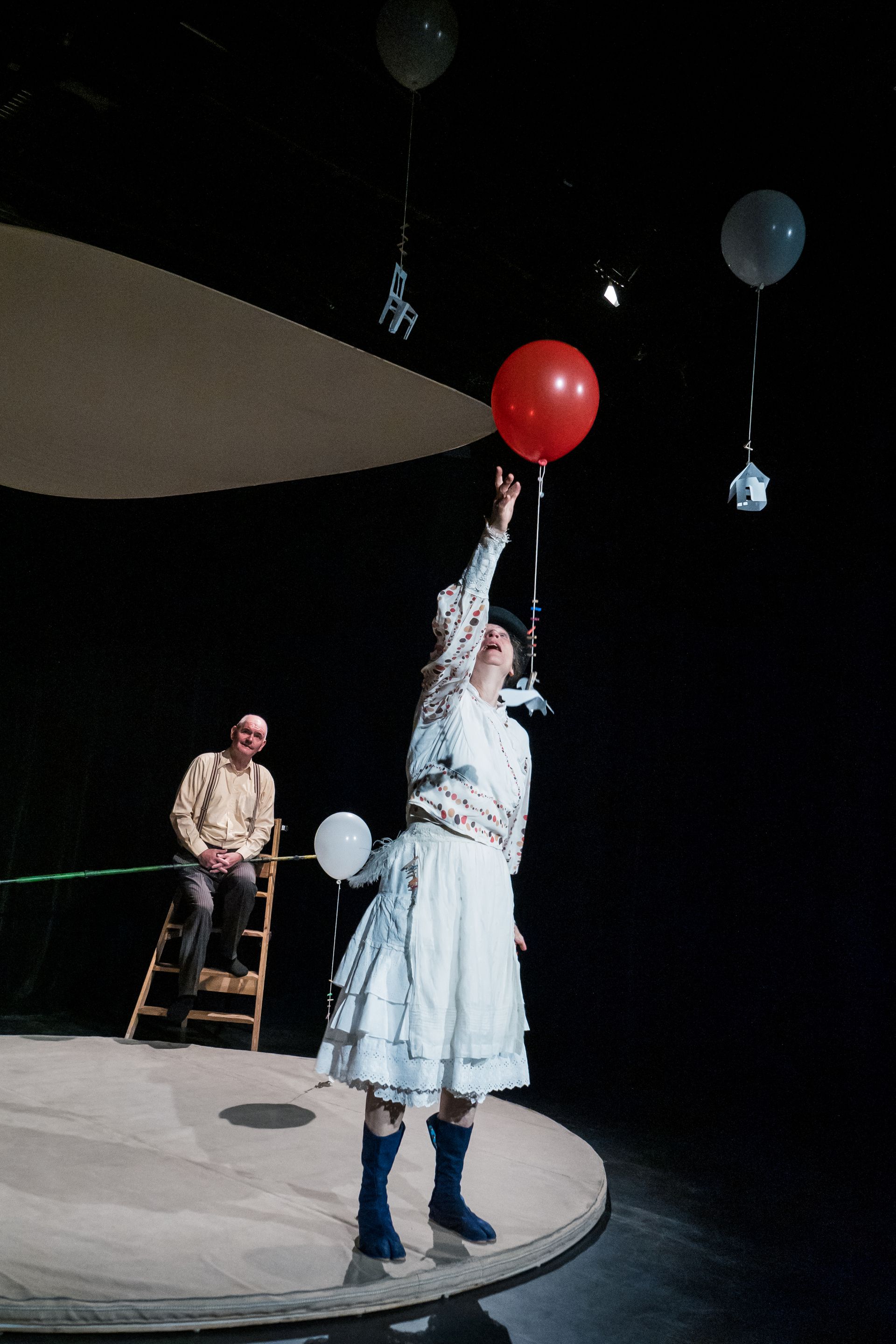 Frau greift auf der Bühne nach einem roten Ballon; Mann sitzt auf einer Leiter. Schwarzer Hintergrund.