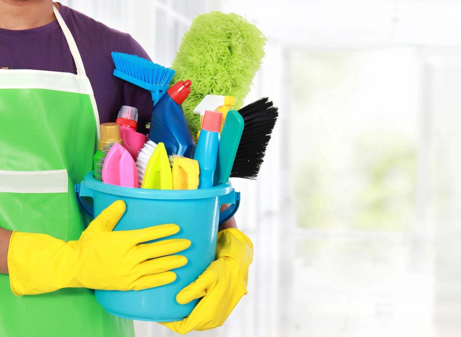 Person wearing gloves and apron, holding a blue bucket filled with cleaning supplies.