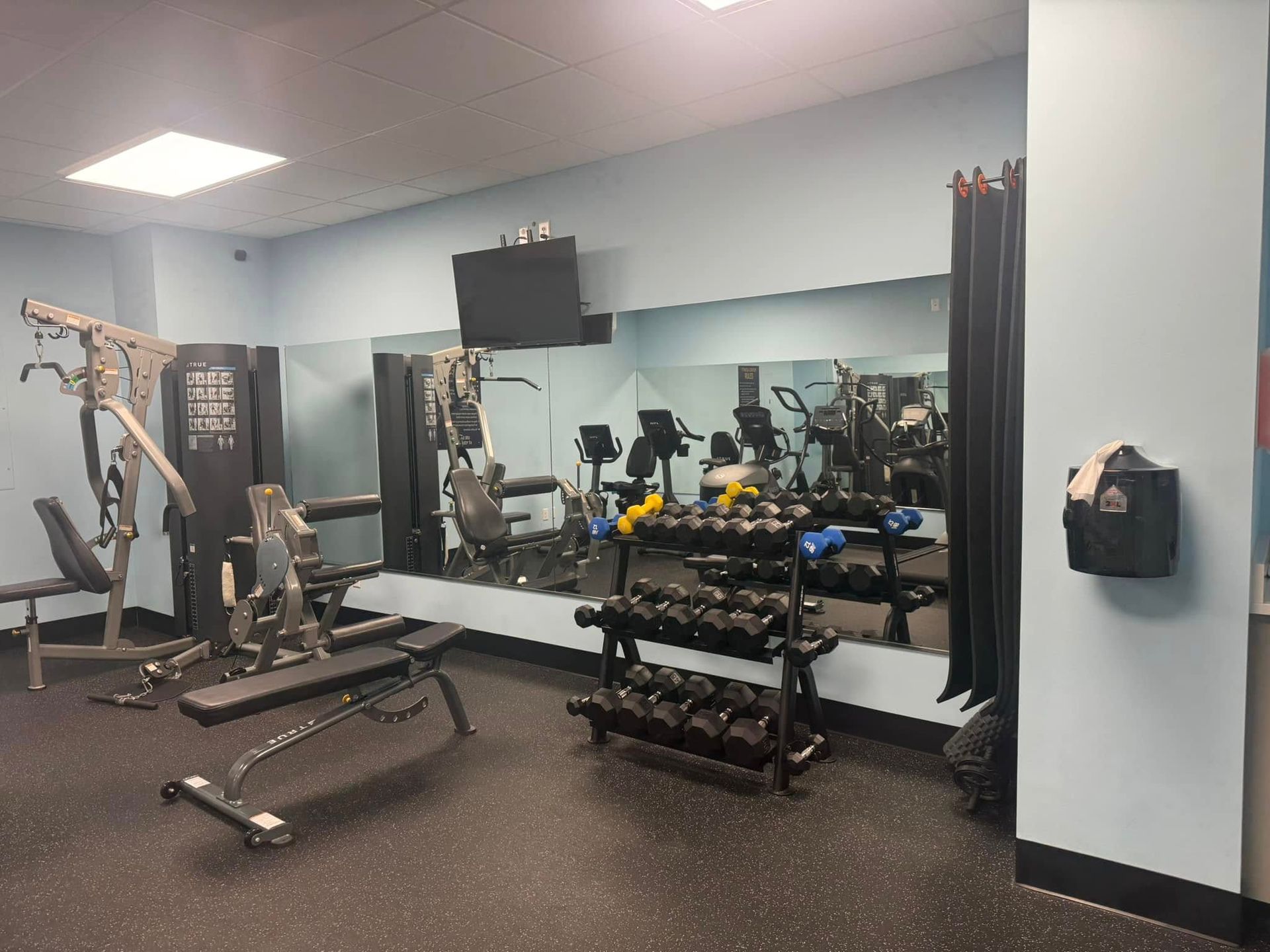 Gym with workout equipment, including weights, weight machines, and a bench. Reflective mirror on the wall.