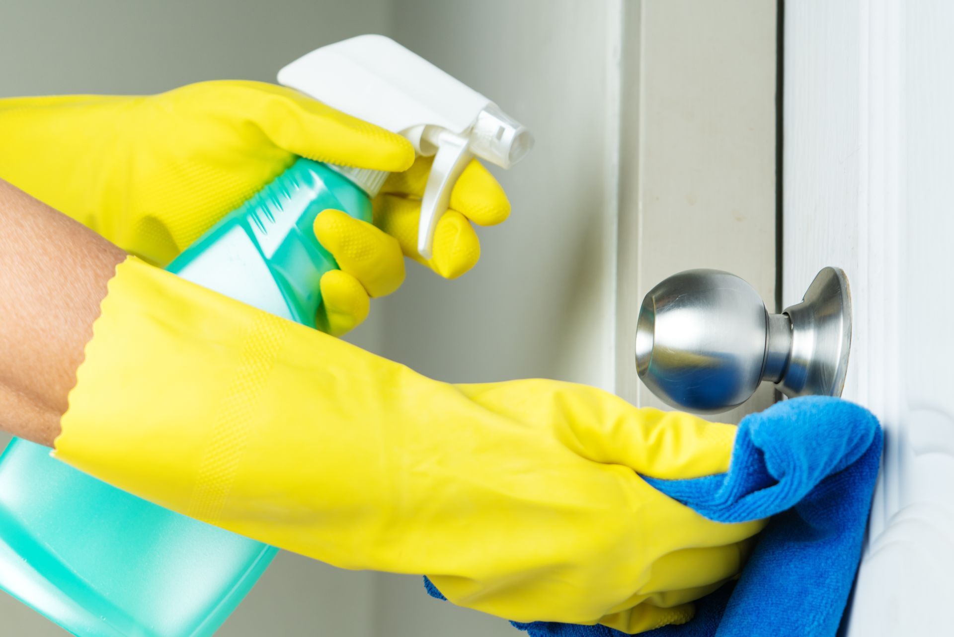 Person in yellow gloves spraying and wiping a doorknob with a blue cloth. Person in yellow gloves spraying and wiping a doorknob with a blue cloth.