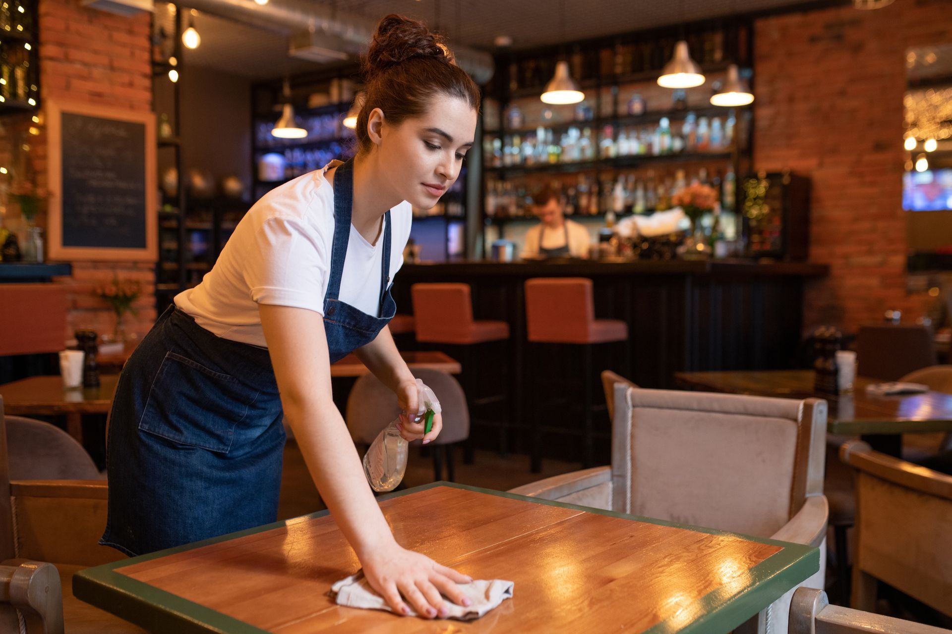 A server in a denim apron cleans a wooden table in a restaurant.