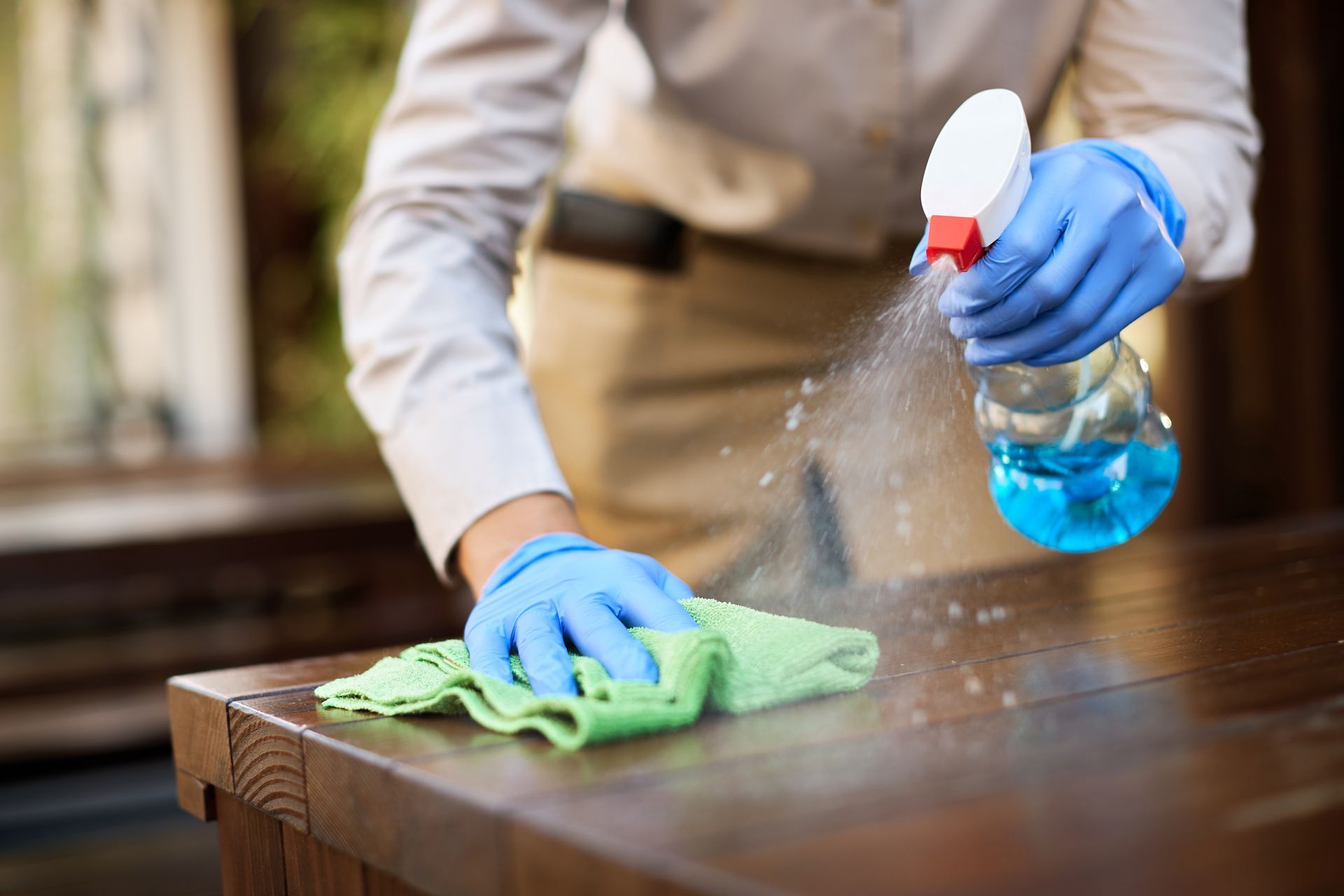 Person wearing gloves spraying and wiping a wooden surface with a green cloth.