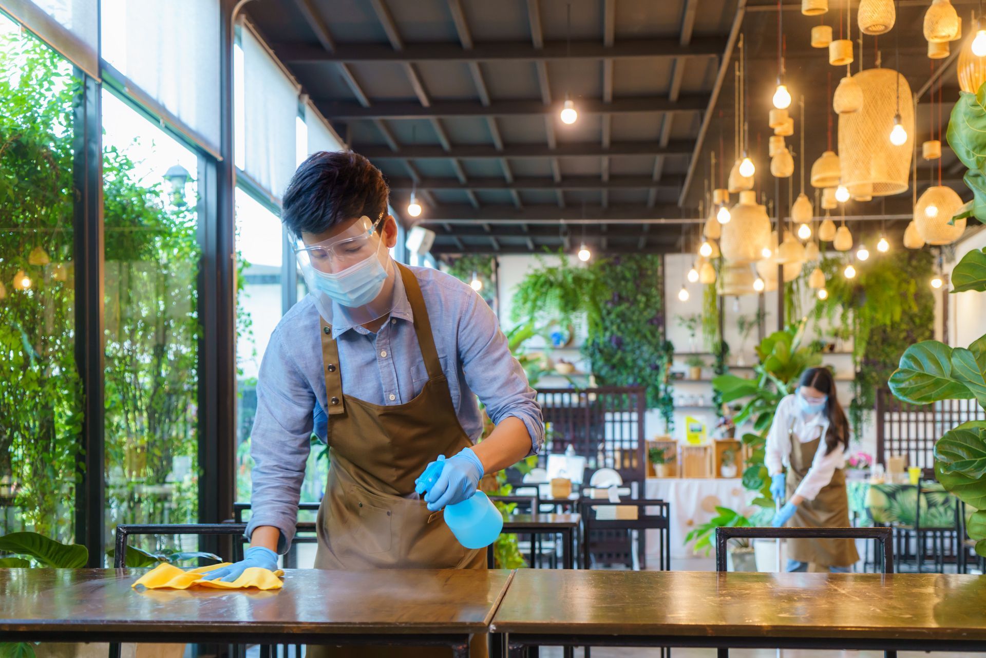Two masked restaurant workers sanitizing tables with spray bottles and cloths.