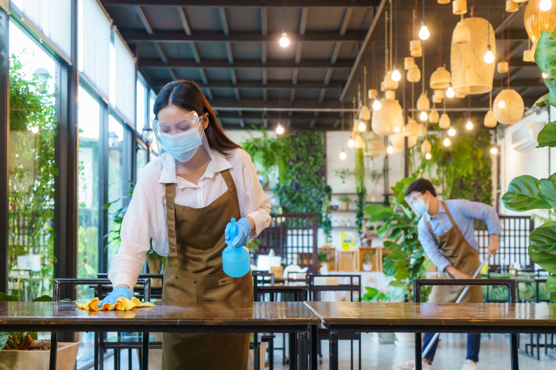 Two people wearing face shields and aprons clean a restaurant.