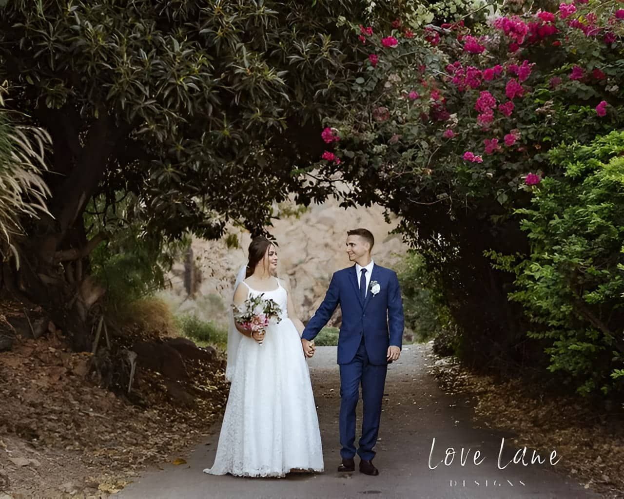 A Bride And Groom Are Walking Down A Path Under A Tree Holding Hands — Lynda Ross Marriage Celebrant In Alice River, QLD