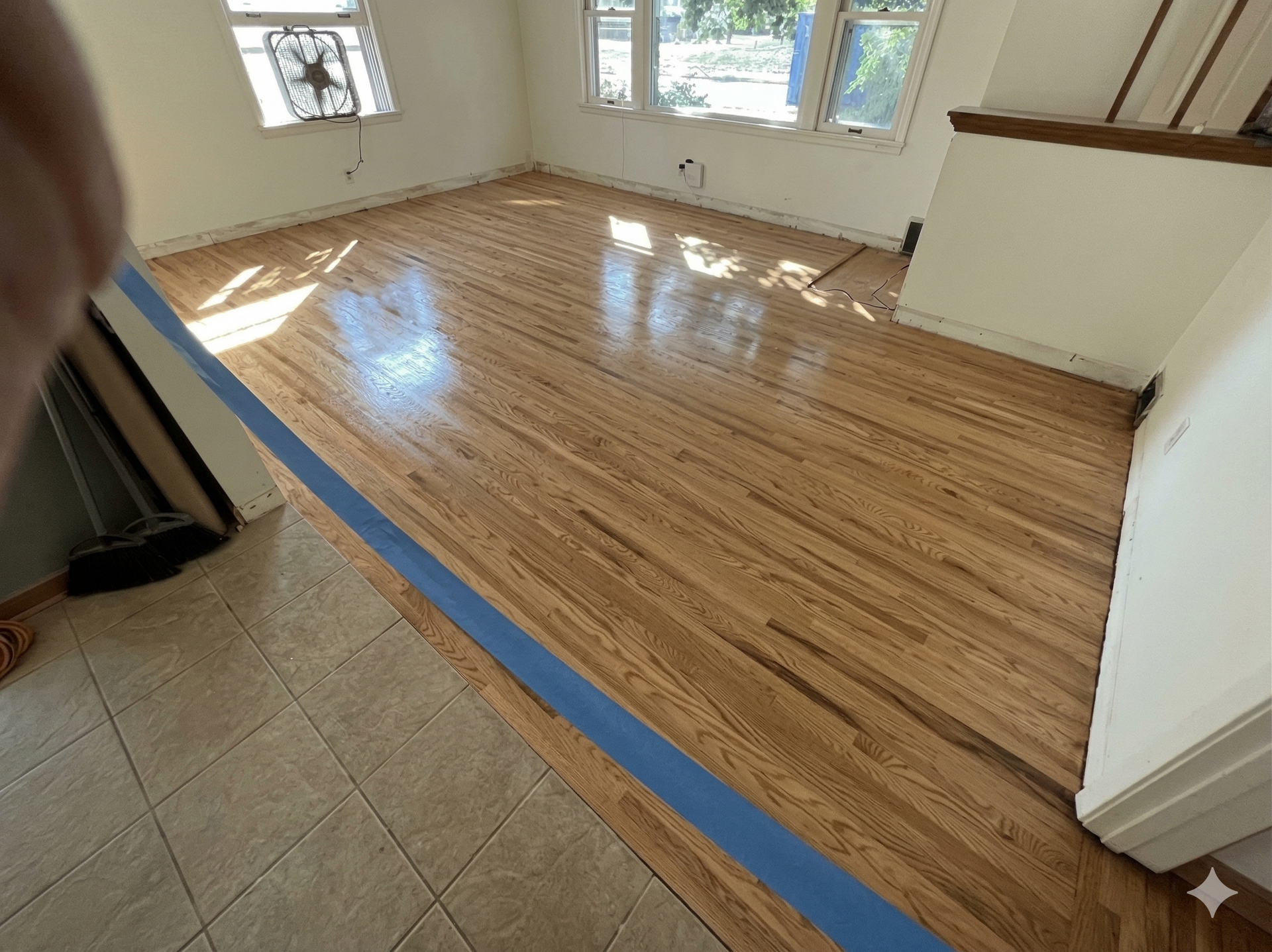 A newly refinished light oak hardwood floor in a room, divided from a tiled area by a strip of blue painter's tape.