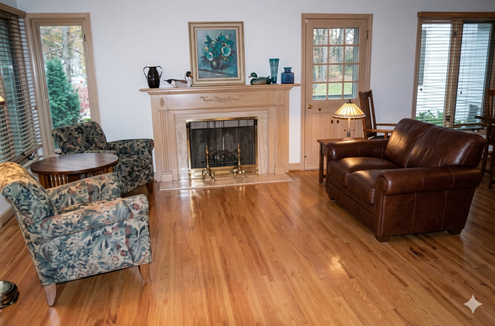 A cozy living room with a fireplace, two patterned armchairs, and a brown leather loveseat on hardwood floors.