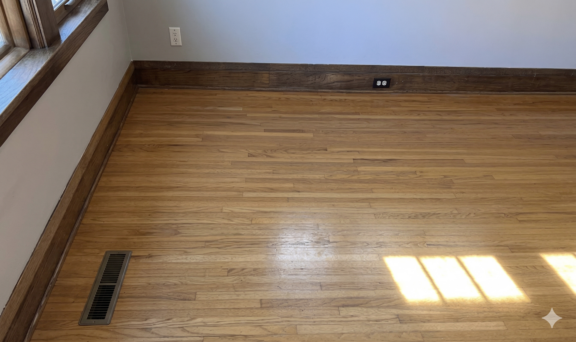A hardwood floor room featuring light-colored planks, a floor vent, dark wood baseboards, and a wall outlet.