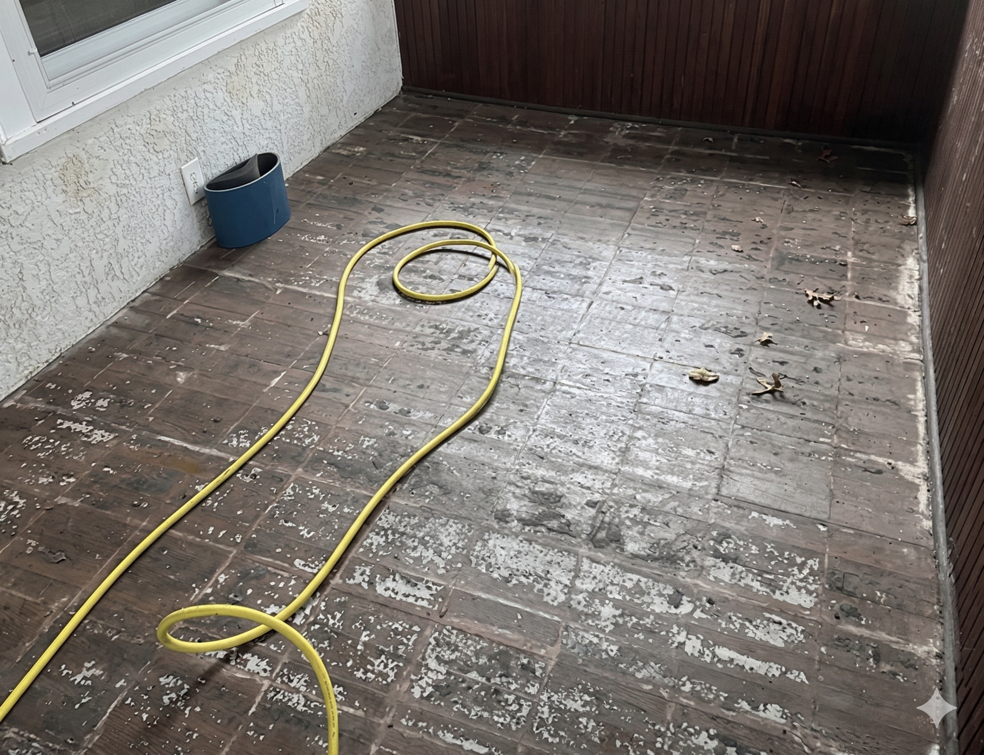 A yellow garden hose lies on a weathered, tiled balcony floor with scattered debris near a wall.