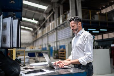 A man in a suit and glasses is standing in a factory with his hands in his pockets.