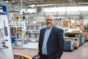 A man in a suit and glasses is standing in a factory with his hands in his pockets.