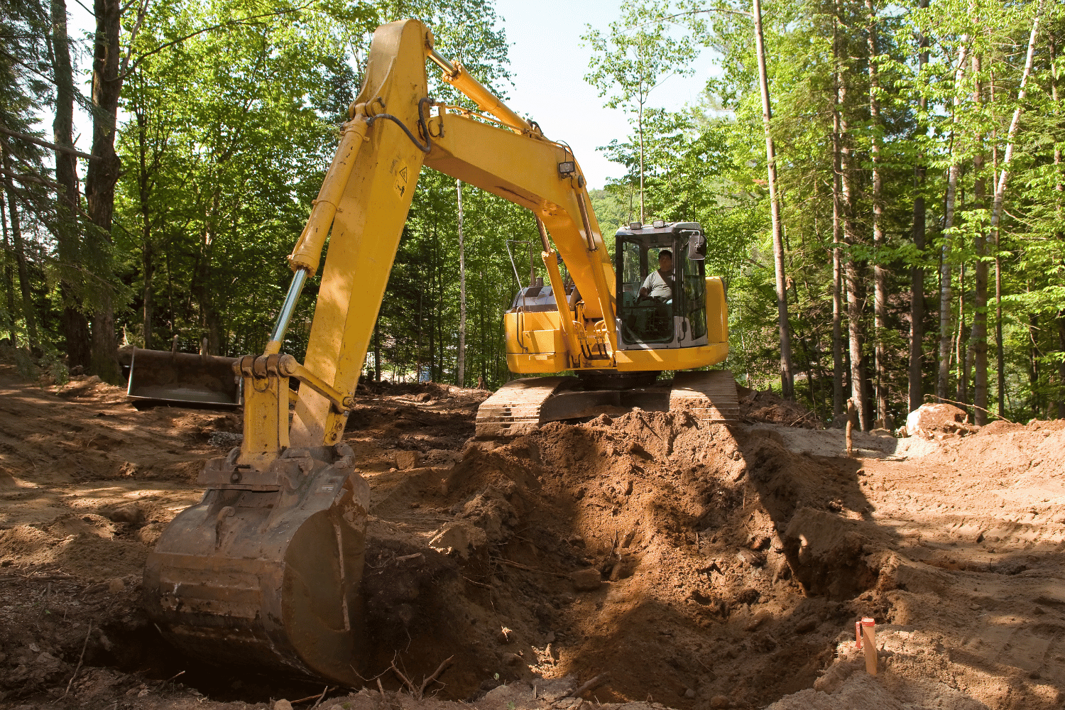 A yellow excavator digs a trench in a wooded area with dirt piles on both sides.