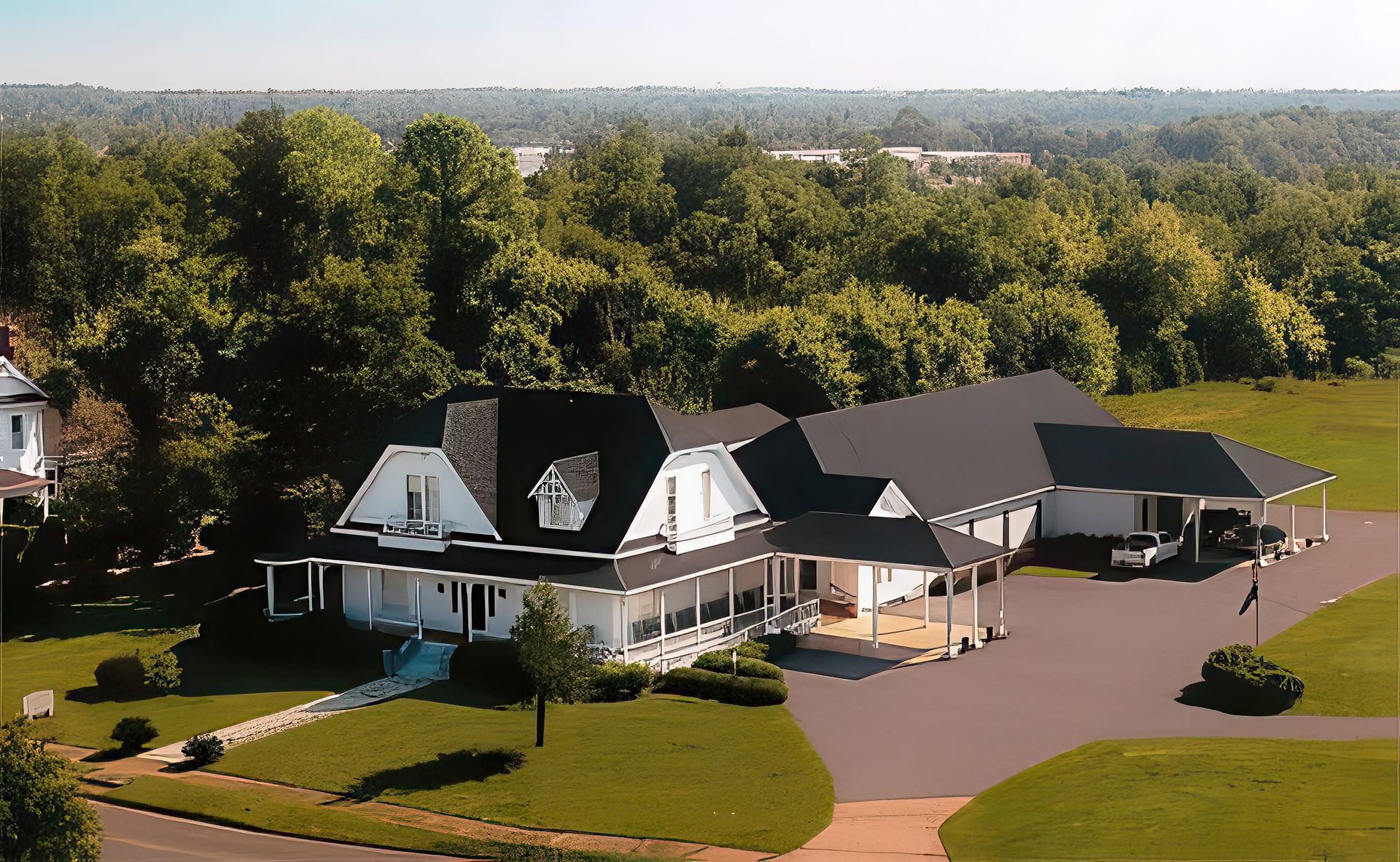 White house with black roof, porch, and long driveway, set against green trees and sky.