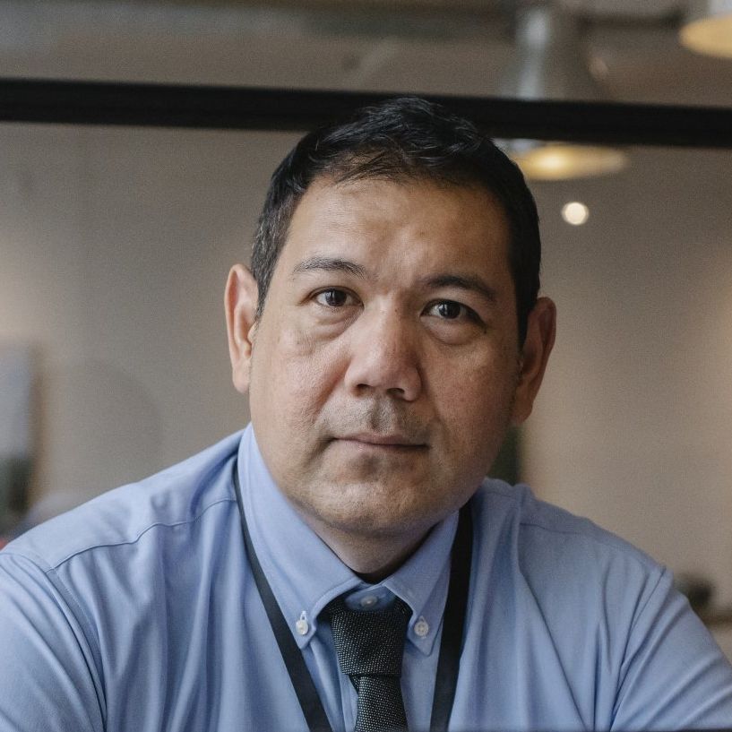 A person wearing a light blue button-up shirt and a dark tie, looking directly at the camera in an indoor setting.