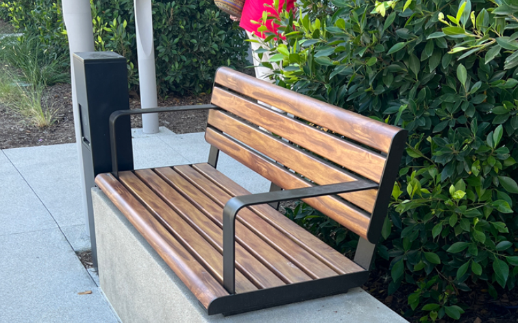 A wooden bench is sitting on a sidewalk next to a bush.