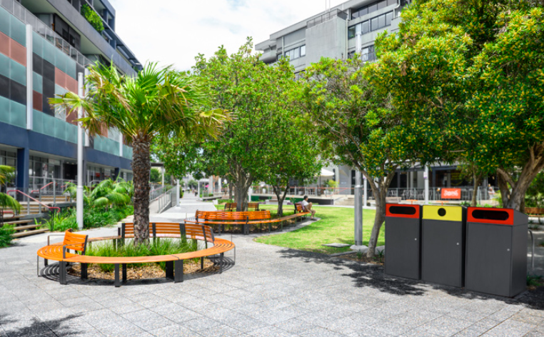 A park with trees , benches , and trash cans in front of a building.