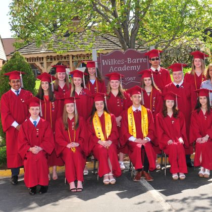 Graduating class of Wilkes-Barre Academy poses in red caps and gowns in front of school sign.