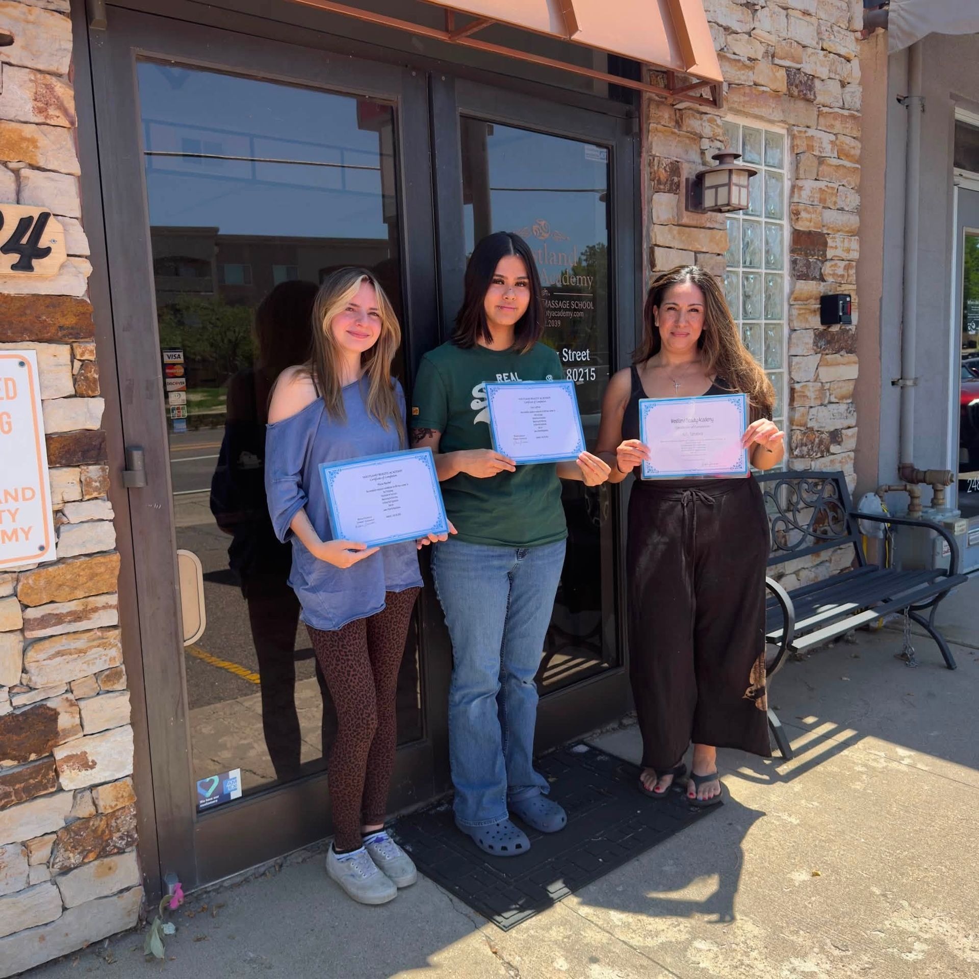Three people holding certificates stand outside a stone building in bright sunlight.