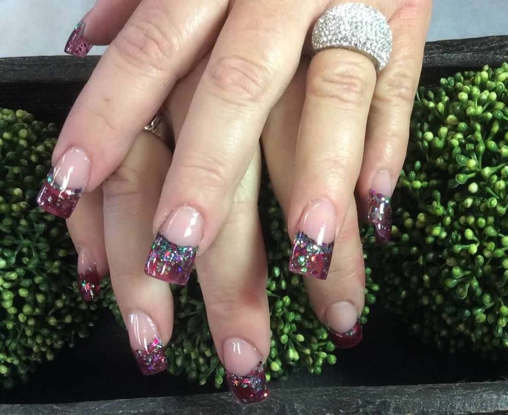 Hands with long glittery red and silver French-tip nails and a rhinestone ring over a textured green-black surface