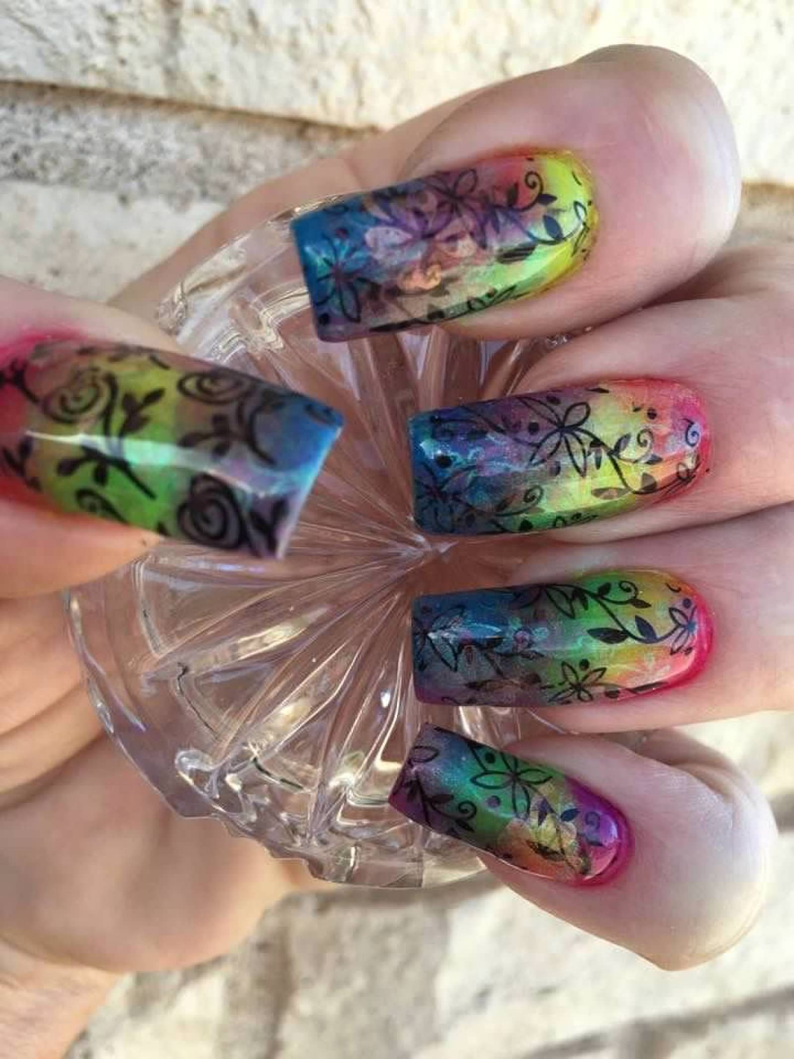 Hand with long rainbow gradient nails and black floral designs holding a clear glass object