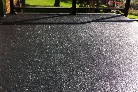 Surface extérieure en béton coloré foncé, à texture granuleuse, éclairée par le soleil, avec des reflets brillants et un environnement extérieur visible en arrière-plan.