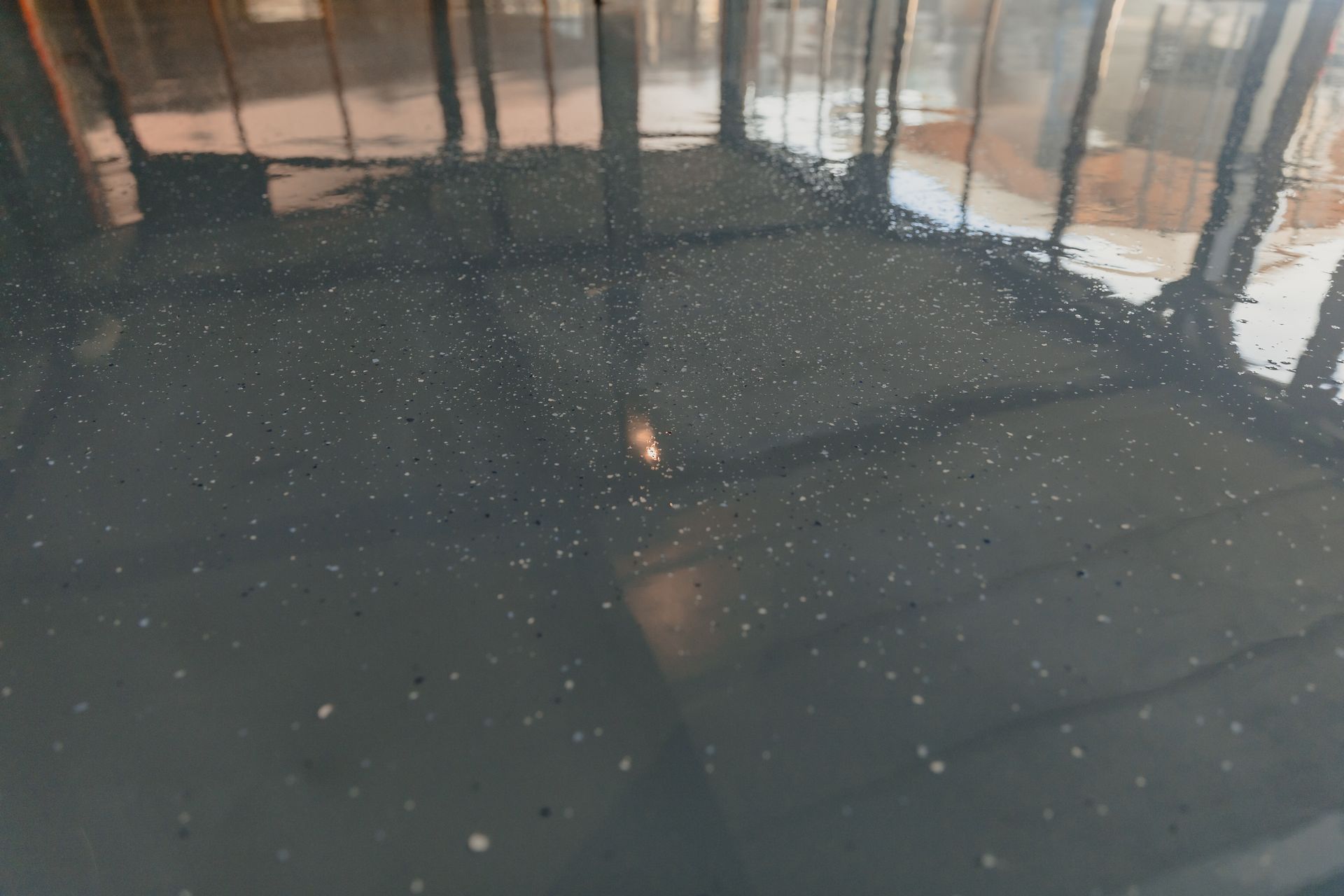 Sol en époxy gris, lisse et humide, avec de fines particules visibles en surface, reflétant les fenêtres et la structure du bâtiment dans une lumière diffuse.