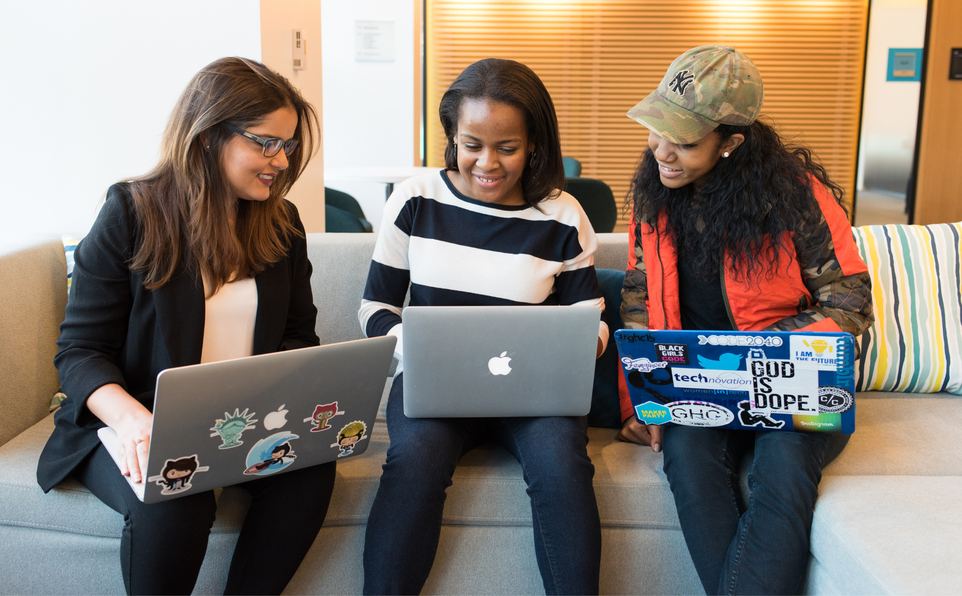 three women are sitting on a couch using laptops .