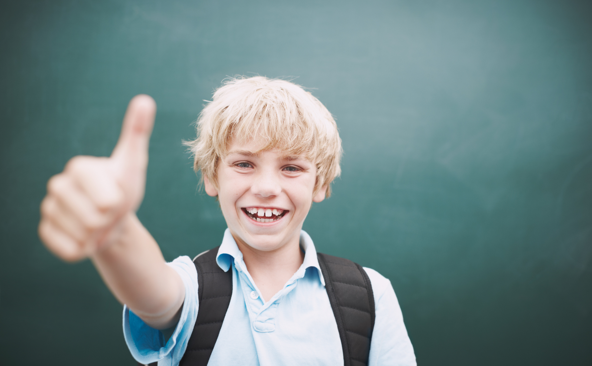 a young boy with a backpack is giving a thumbs up in front of a blackboard .