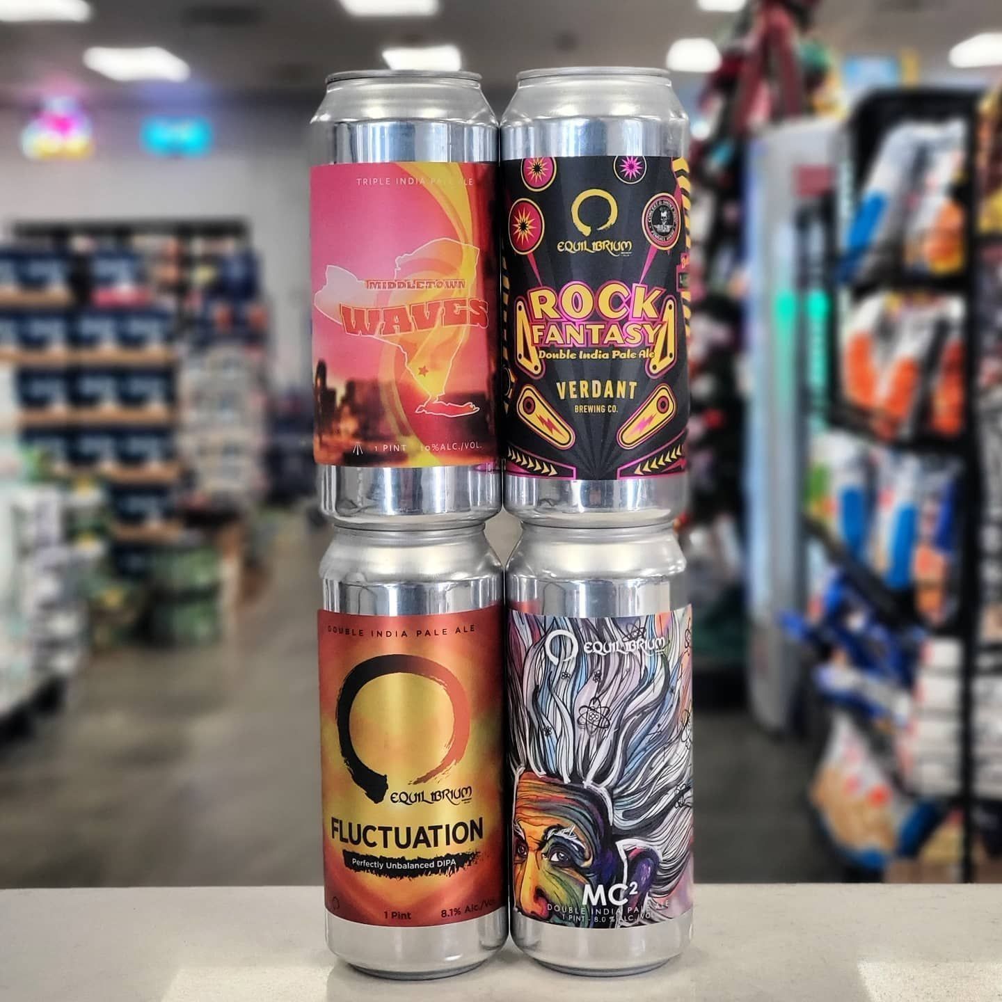 Four cans of craft beer stacked vertically against a blurry store backdrop. The cans have colorful labels.