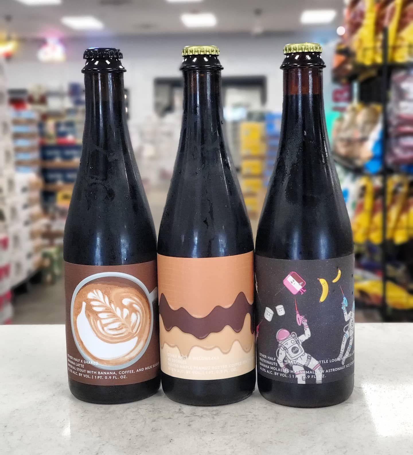 Three dark beer bottles with colorful labels on a store counter, depicting coffee art, layers, and astronauts.
