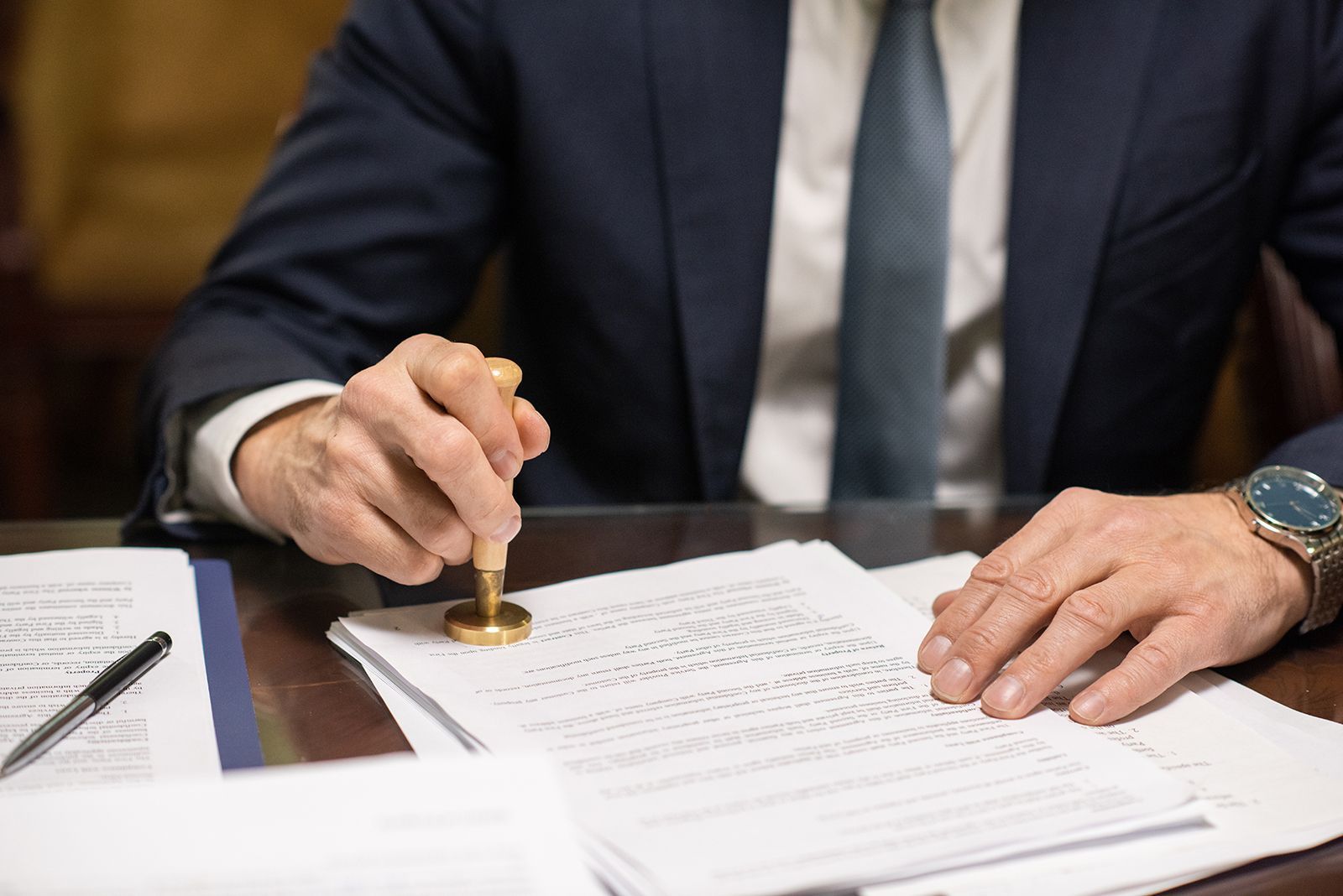 Una persona con traje sella un documento con un sello, probablemente para su aprobación oficial, en un escritorio.