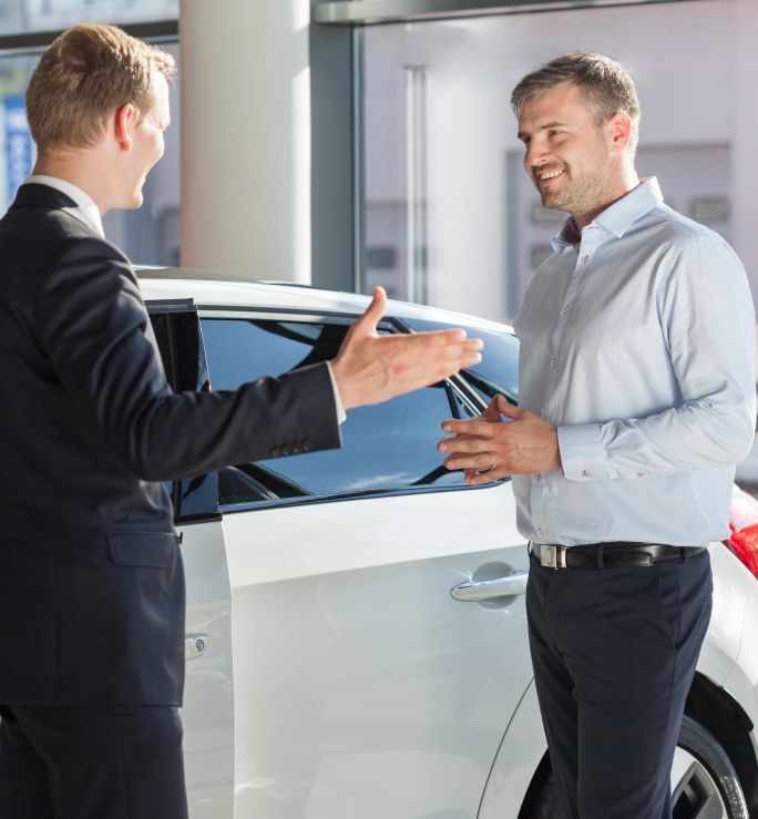 Two men are talking in front of a white car