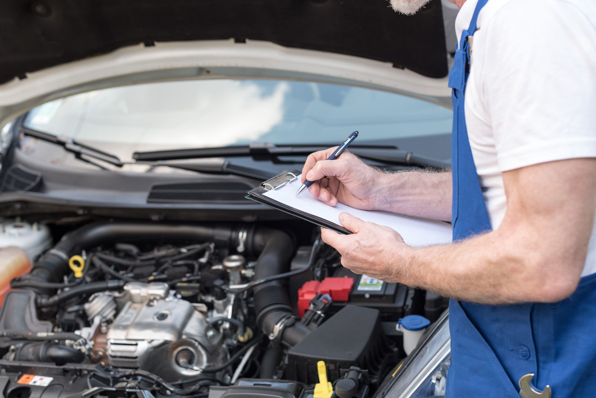 Mechanic inspecting a car engine with a clipboard under the open hood Mechanic inspecting a car engine with a clipboard under the open hood