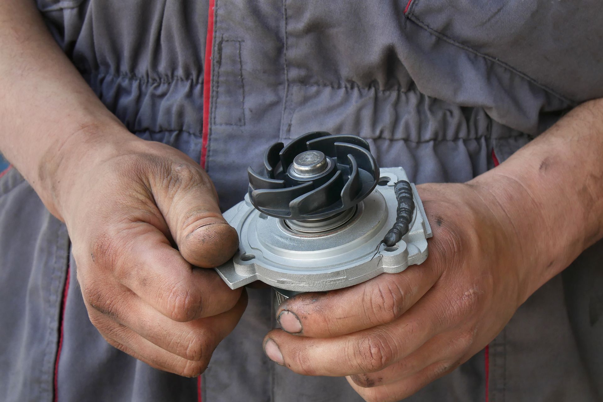 Hands holding a small metal mechanical part with a black impeller, against a gray work jacket background