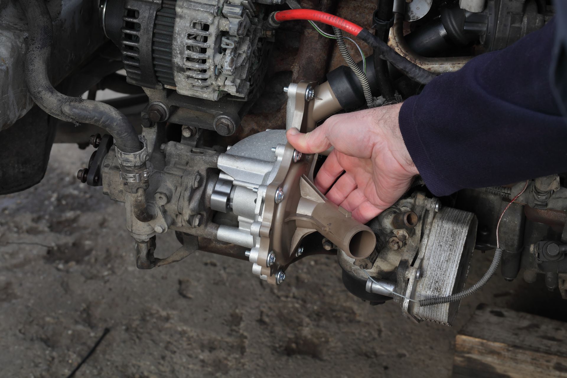 Hand holding a silver automotive part in a dirty engine bay, with exposed wires and components.