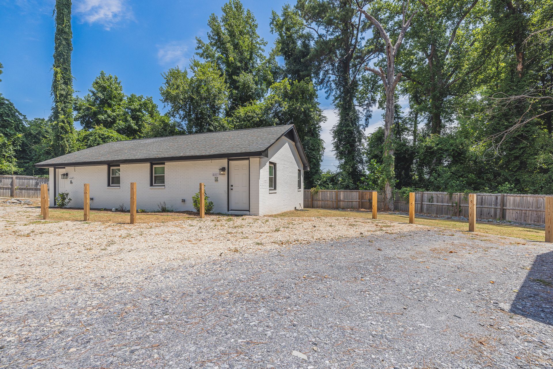 A small white house is sitting in the middle of a gravel lot surrounded by trees.