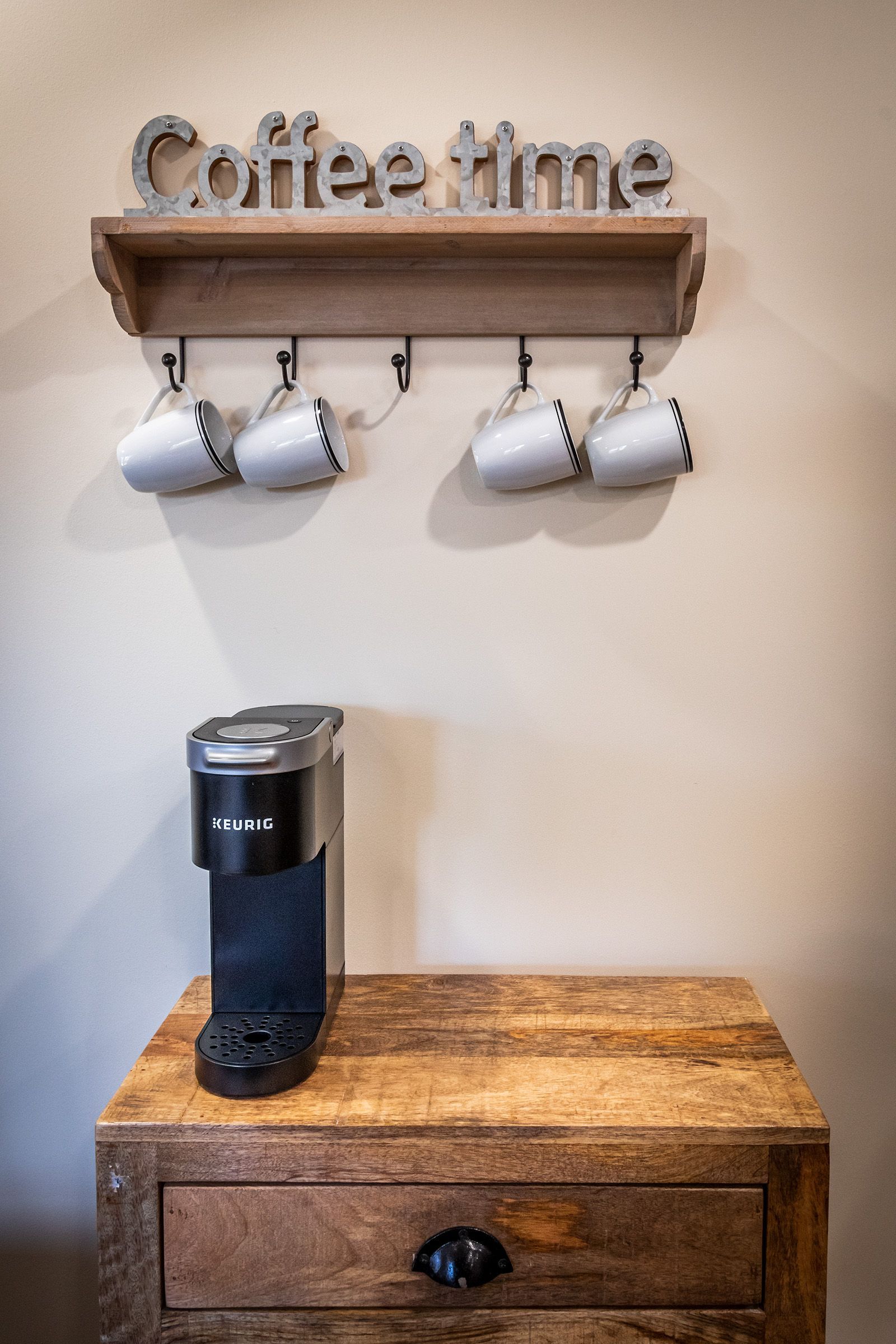 A coffee maker is sitting on a wooden table next to a shelf with cups hanging from it.