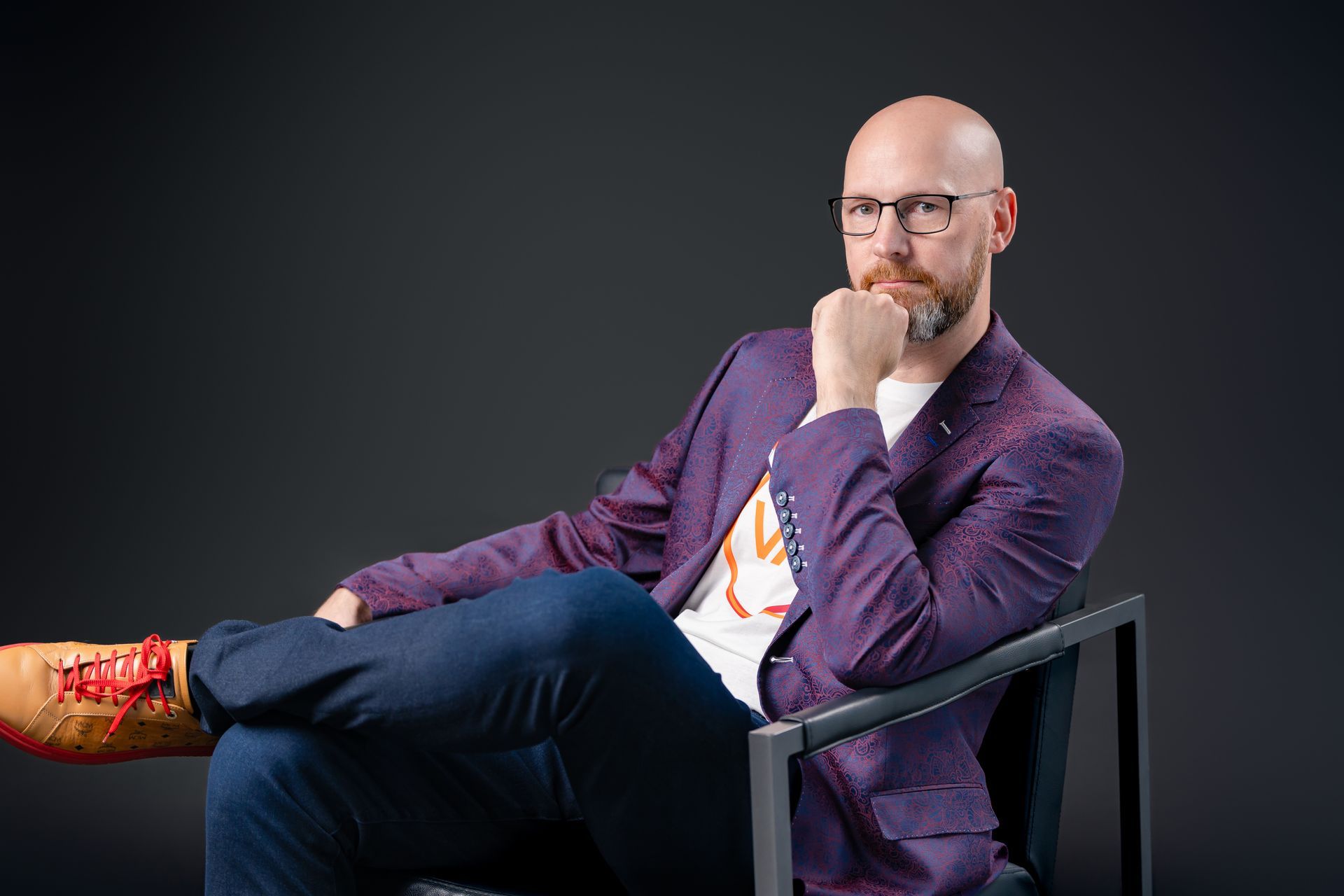 A man in a purple jacket and glasses is sitting in a chair with his legs crossed.