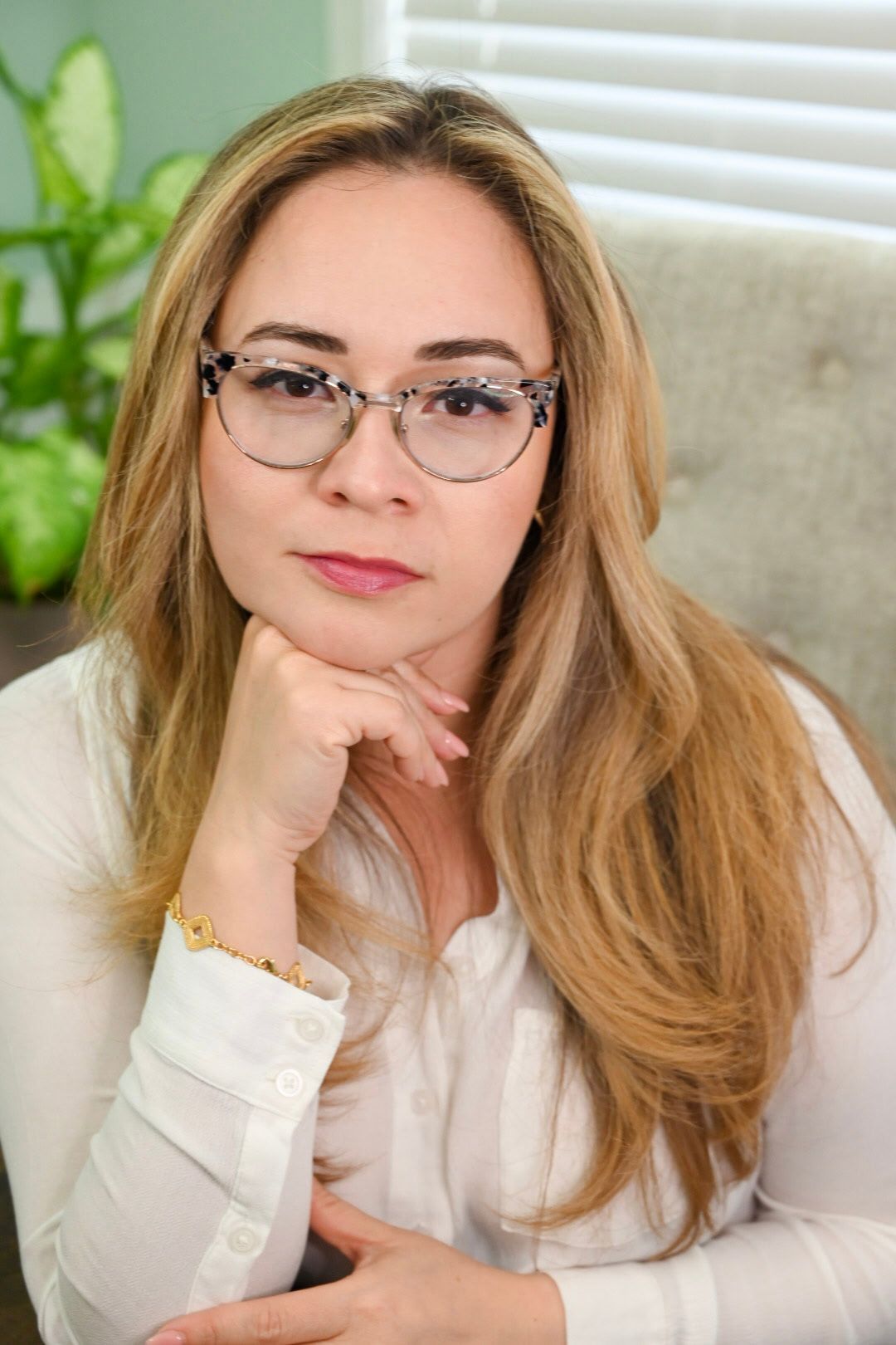 A woman wearing glasses and a white shirt is sitting in a chair with her hand on her chin.