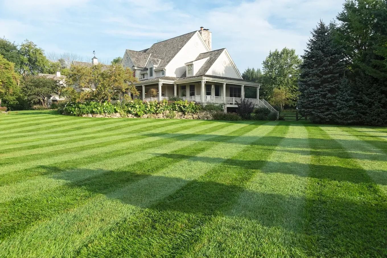 Lawn with striped mowing pattern in front of a large house with a porch, under a sunny sky.