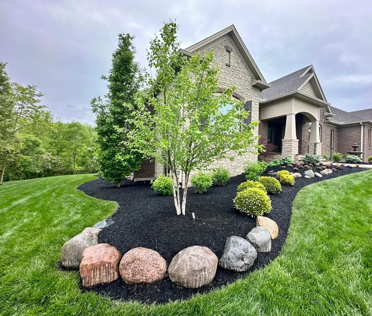 A house with a landscaped front yard, featuring a bed with dark mulch, trees, and stones.