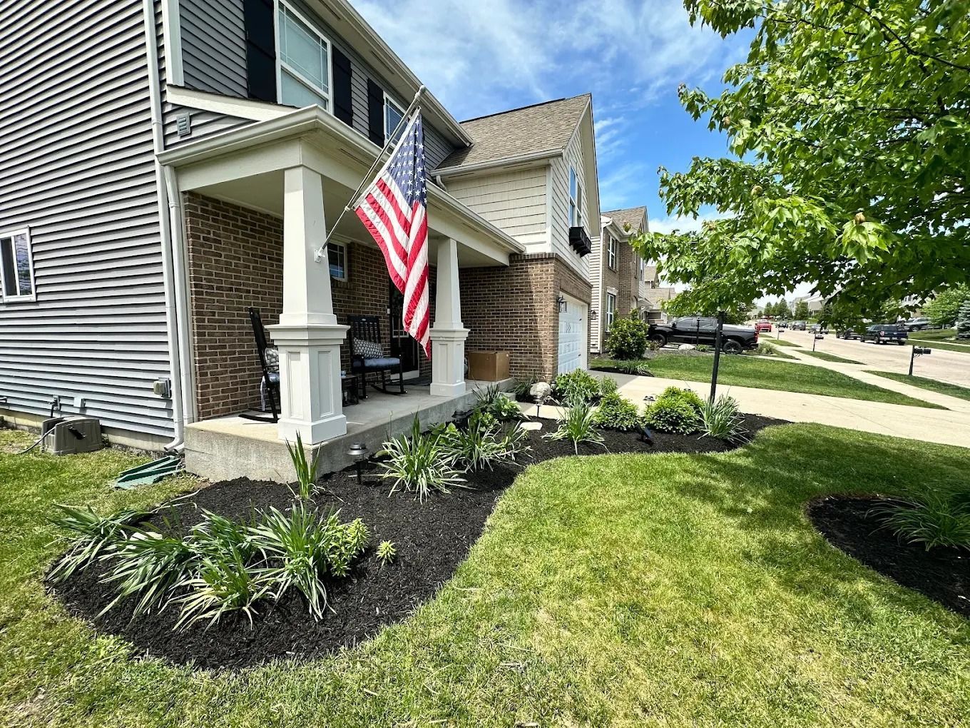 A house with a porch, American flag, and garden, on a sunny day.