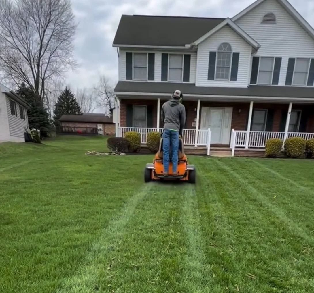 Man mowing lawn in front of two-story house with white trim and green shutters on a cloudy day.