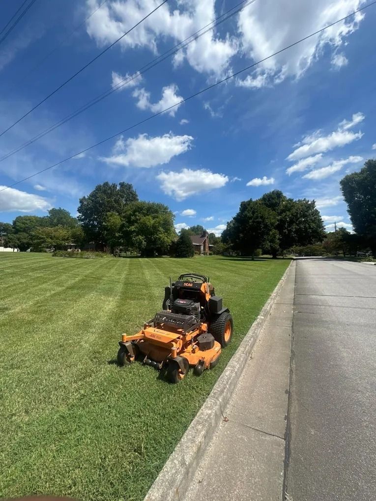Orange riding lawnmower on green grass, next to a road, under a blue sky with clouds.