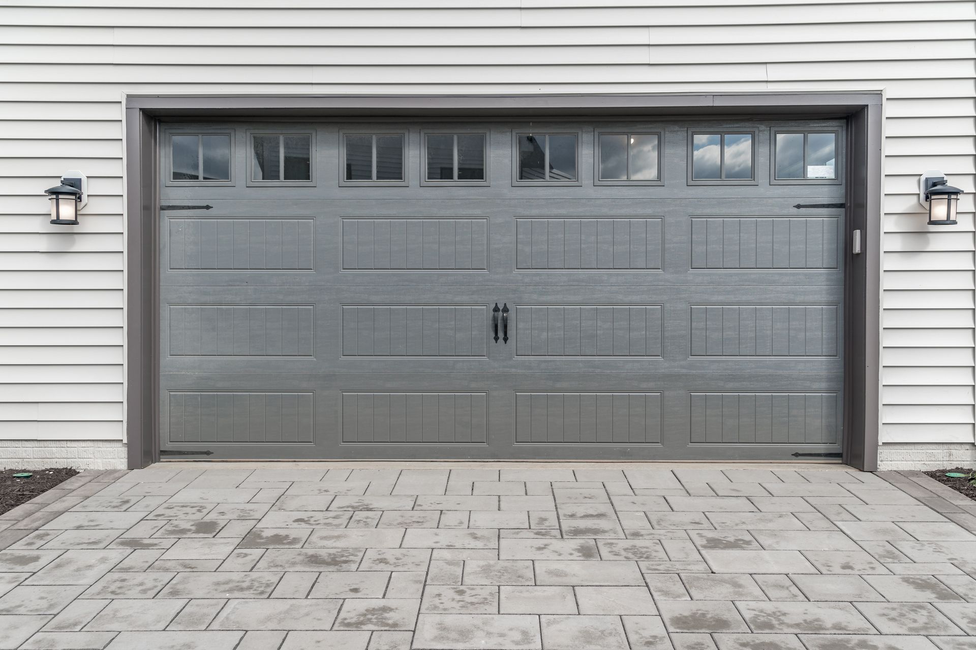 Porte de garage grise avec fenêtres supérieures et deux appliques murales, située devant une allée en briques.