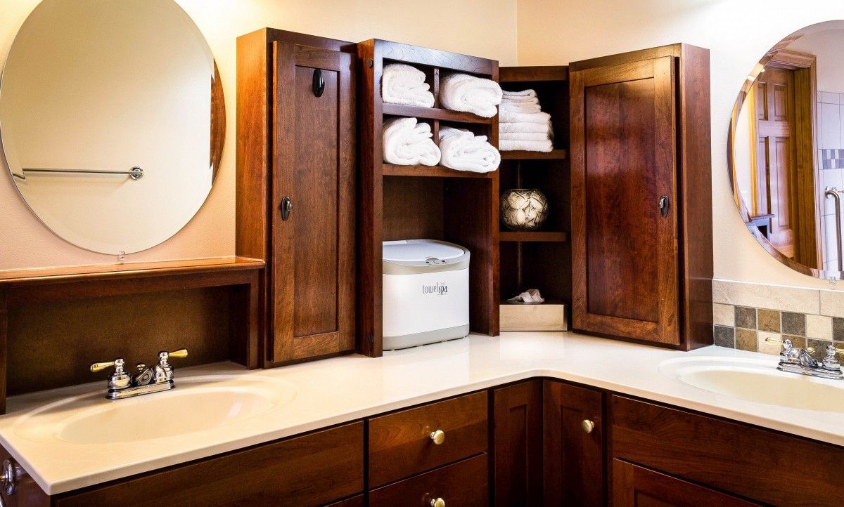Salle de bains avec meubles en bois foncé, comptoir blanc, deux lavabos et une armoire de rangement pour les serviettes.