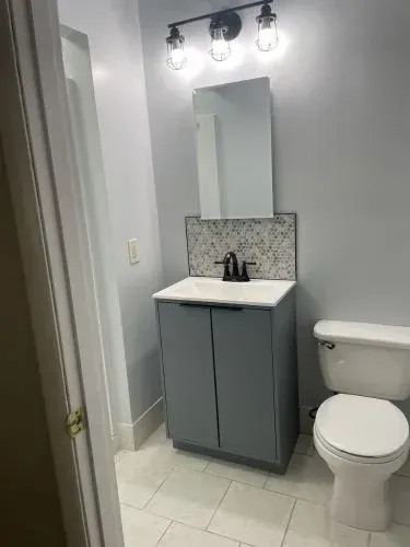 Small bathroom with gray vanity, white toilet, tiled floor, and wall mirror under lights.