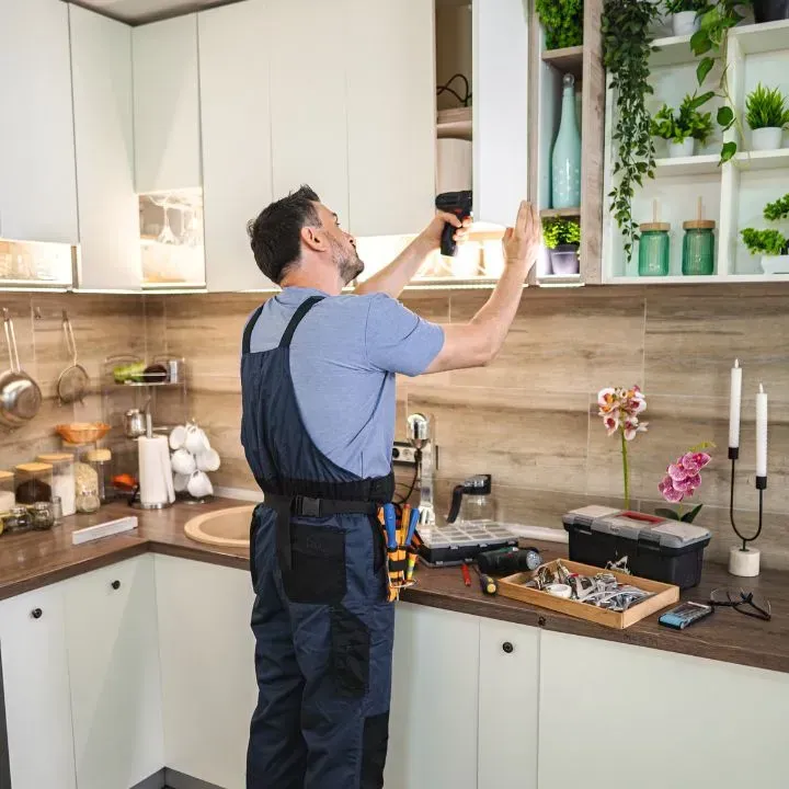 Handyman in blue overalls, working on kitchen cabinet with a drill.