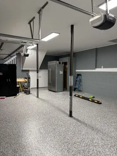 Spacious garage with gray speckled floor, metal poles, overhead equipment, cabinets, and a snowboard on the floor